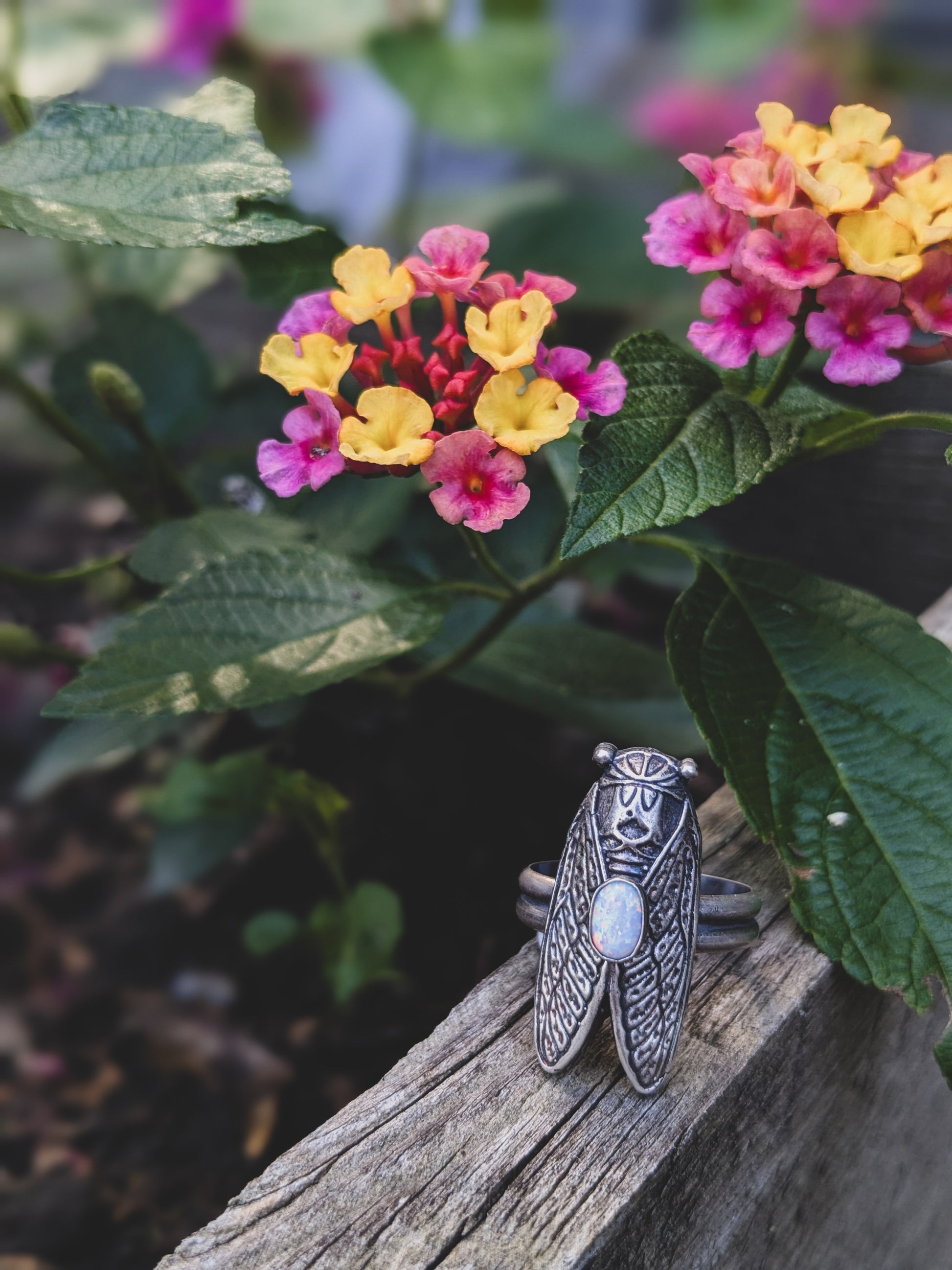 Sterling silver cicada with an opal stone sits on a wood under orange and pink flowers
